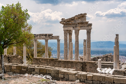 Temple of Trajan ruins, Acropolis of Pergamon, Bergama, Turkey. Ancient Roman columns, stone walls, and green trees under a cloudy sky, overlooking the city.