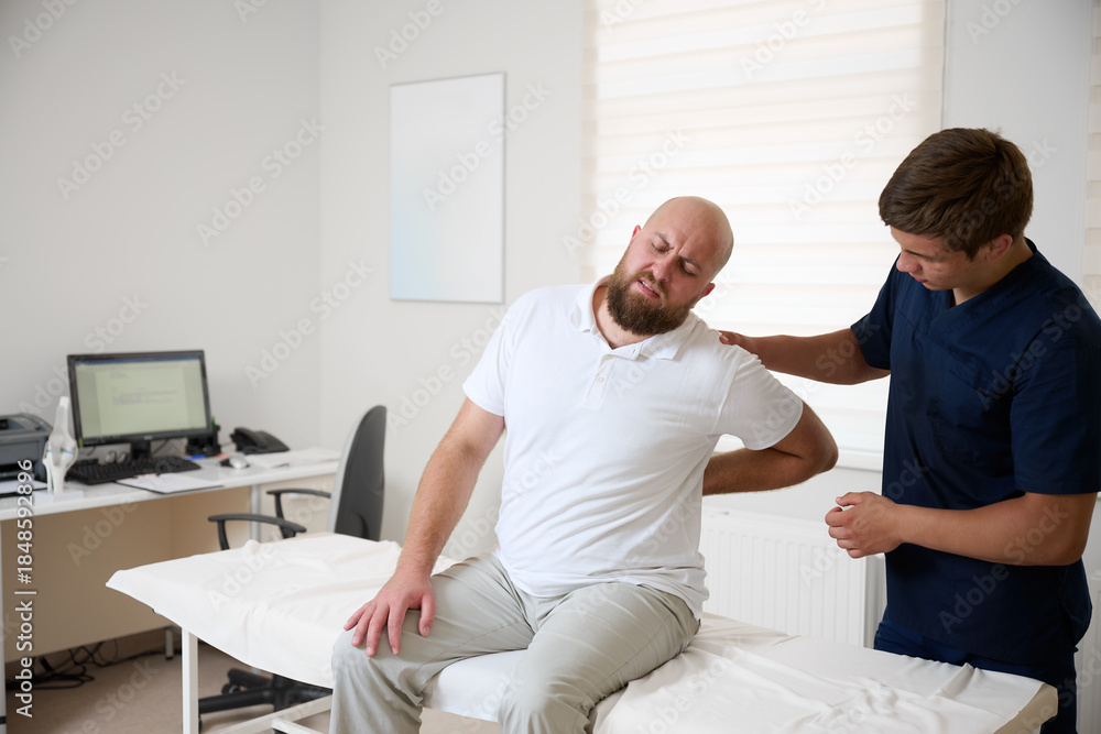 Fototapeta premium Man suffering from back pain sitting on examination table in physiotherapy clinic. Concept of spine problems, rehabilitation, and medical consultation.