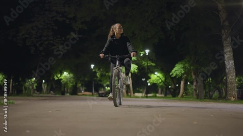 Wallpaper Mural Solo Woman Pedals Beneath Tranquil Streetlights, Serene Young Woman Cycling Through Quiet Park At Night, Calm Female Cyclist Traverses Silent Park Illuminated By Gentle Streetlamps In Dark Torontodigital.ca