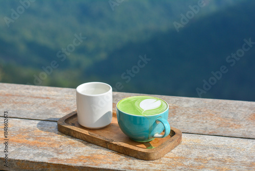 Hot matcha green tea in a blue cup, served with hot water, on a wooden table and blurry nature background.