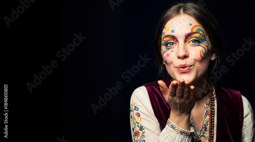 Woman with rainbow face paint and a white vest. She is blowing a