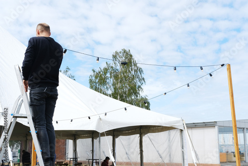Worker on ladder prepares a large white tent for celebration, hanging decorative lanterns and string lights, creating a festive atmosphere for outdoor event