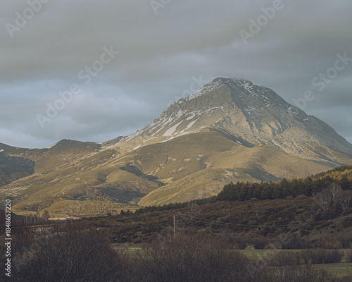 View of mountainous landscape with golden sunlight illuminating the slopes and a dusting of snow on the peak under a cloudy sky, Riano, Castilla y León, Spain.