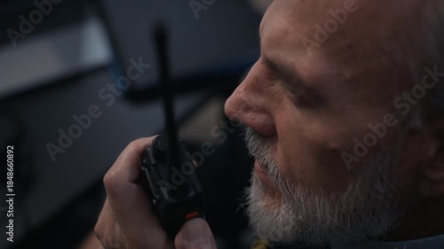 Close view of adult Caucasian male security officer speaking into two way radio while sitting at desk in front of surveillance screens in dark control room
