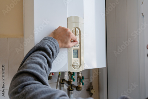 Man’s hand regulating the temperature dial of a wall-mounted boiler with exposed plumbing, illustrating residential heating control and energy efficiency.