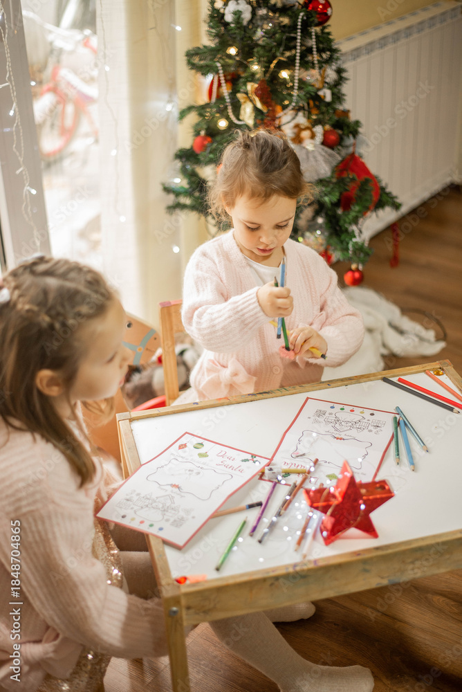 Fototapeta premium Cozy Christmas moment of two adorable little sisters drawing together at a small wooden table near a decorated Christmas tree, warm fairy lights in the background, soft pastel outfits, authentic famil