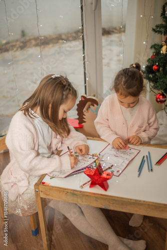 Cozy Christmas moment of two adorable little sisters drawing together at a small wooden table near a decorated Christmas tree, warm fairy lights in the background, soft pastel outfits, authentic famil