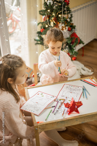 Cozy Christmas moment of two adorable little sisters drawing together at a small wooden table near a decorated Christmas tree, warm fairy lights in the background, soft pastel outfits, authentic famil