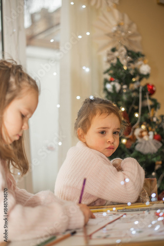 Cozy Christmas moment of two adorable little sisters drawing together at a small wooden table near a decorated Christmas tree, warm fairy lights in the background, soft pastel outfits, authentic famil