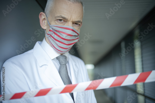 Senior doctor white coat wearing striped face mask looks concerned while standing behind red barrier tape hospital corridor, serious protective