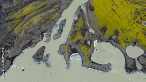 Aerial view of the stark, contrasting landscape where the glacial river meets the moss-covered earth, creating a tapestry of gray, green, and pale blue hues, Solheimajokull, Myrdalshreppur, Iceland.