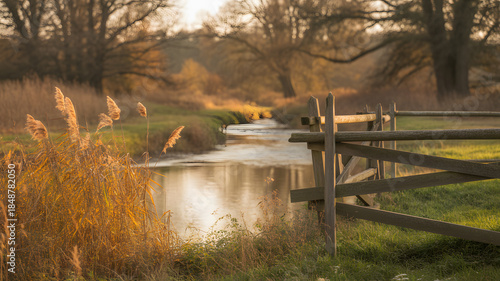 Sunlit river flows through countryside scene with autumn reeds and a rustic wooden fence in foreground, capturing a tranquil landscape