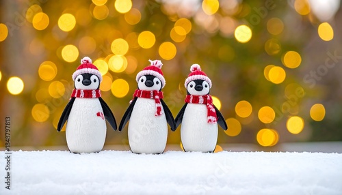 Three plush penguins wear red hats and scarves standing in the snow with blurred lights in the background