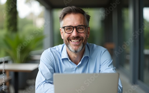 Happy confident mature professional business man hybrid working looking at camera. Smiling middle aged businessman wearing glasses sitting at outdoor office table using laptop. Portrait. Copy space.