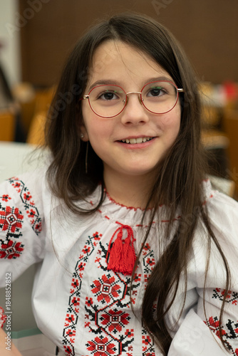 Vertical photo of smiling schoolgirl in glasses wearing vyshyvanka sitting in classroom, education concept