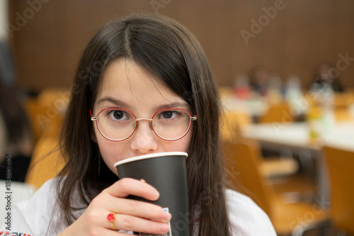 Schoolgirl in glasses drinking from paper cup while sitting in classroom, childhood education concept