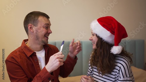 Happy wife surprising her husband with a positive pregnancy test as a christmas gift, making him overjoyed as they celebrate the wonderful news with a loving kiss in their bedroom