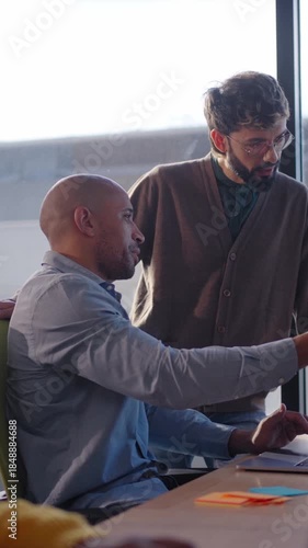 Vertical. Diverse team gathers around a screen for a project meeting, exchanging ideas and planning next steps in a modern office environment with daylight and city skyline in the background.