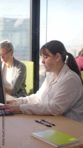 Vertical. Young caucasian woman manager works at her laptop in a bright corporate office with city views, focused on typing and analysis during a productive workday in a modern workspace.