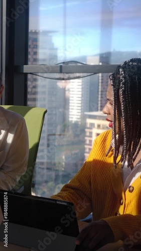 Vertical. Two female colleagues review a project at a desk, pointing at a monitor and laptop in a office with city views, conveying teamwork, guidance, and productive collaboration during a meeting.