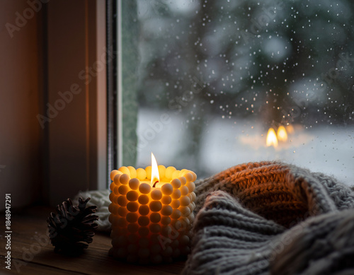 A close-up of a handcrafted wax candle flickering next to a snowy window. Ai