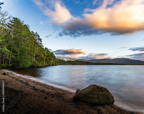 View of a serene loch mirroring the pastel sky, fringed by dark forest and sandy beach, with a mossy rock adding texture, Caledonian Forest, Cairngorms National Park, Scotland.