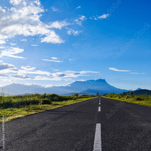 A scenic perspective of the Tempasuk Plains, where a long highway points directly toward the granite massif of Mount Kinabalu, a UNESCO World Heritage site.
