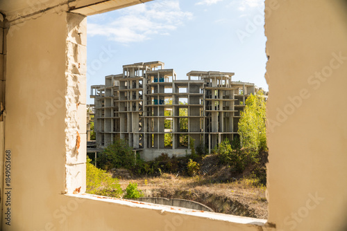 View of an unfinished concrete structure stands amidst greenery and a pit, framed by a weathered window, hinting at abandonment, Costa del croco, Tsarevo, Bulgaria.