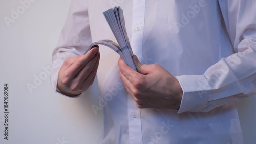 Young man in white shirt on white background flipping through thick stack of dollar bills