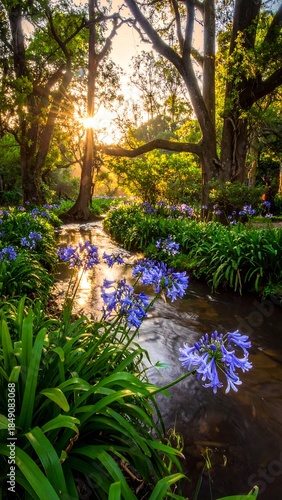 Sunlit stream flows amidst vibrant blue flowers & lush green foliage beneath tall, branching trees in a tranquil setting