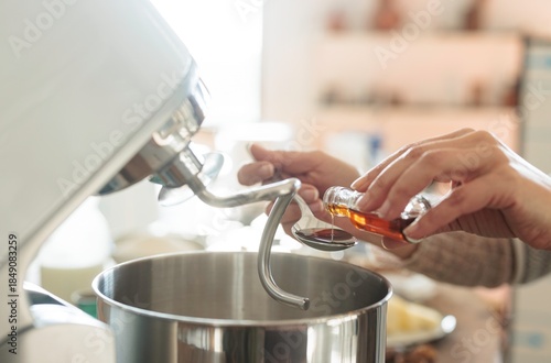 A person is carefully adding vanilla extract to a spoon, poised over a large metal mixing bowl attached to a stand mixer. The kitchen appears bright and cheery