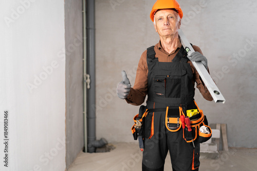 Tableau sur toile Positive senior builder wearing tool belt holding spirit level and showing thumbs up gesture during home renovation