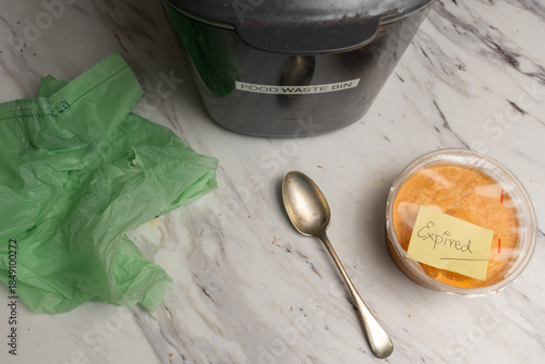 Food waste bin, biodegradable bag, spoon, container of expired food marked with a sticky note. Top view.