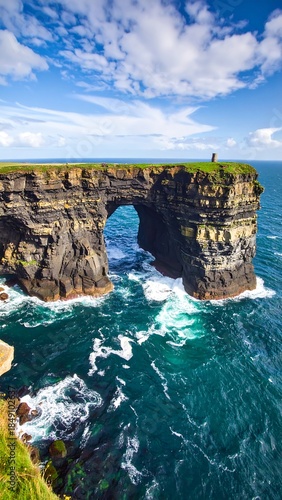 Coastal archway featuring layered rock formations and turbulent turquoise water under a blue sky with scattered clouds