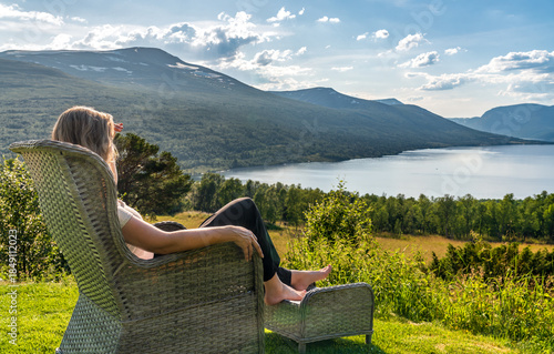 A woman sits in a garden chair with her feet up, looking at Gjevillvatnet lake and mountains in Oppdal, Trøndelag Norway during summer. She enjoys the view and the sun