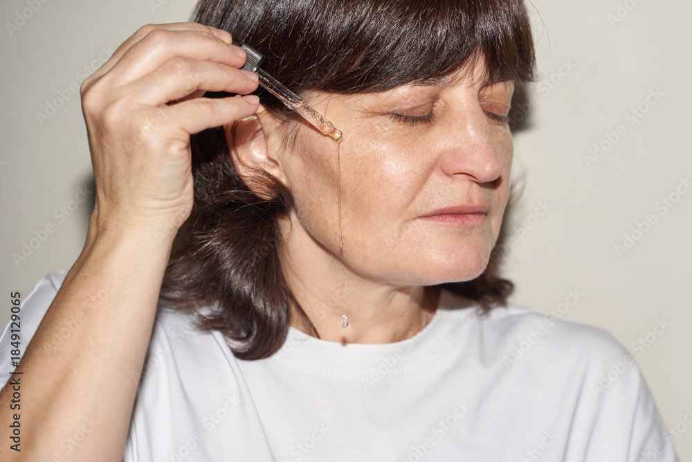 Fototapeta premium Mature caucasian woman applying serum on face for skincare routine and self-care.