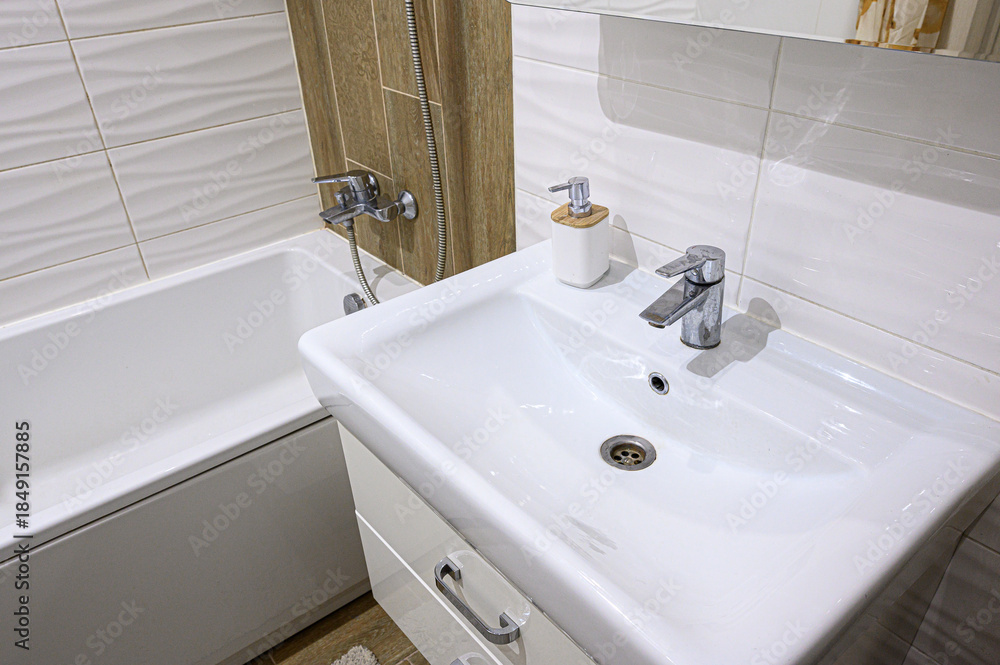 Fototapeta premium modern bathroom features a white ceramic sink with a chrome faucet, a soap dispenser, a white bathtub with a chrome shower tap, light-toned tiles, and wooden-style wall panels