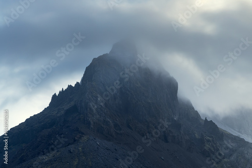 Dark, rugged mountain peak rising into low clouds and mist, creating a moody and dramatic atmosphere. Remote highland landscape emphasizing raw geology, isolation, and powerful natural forms.