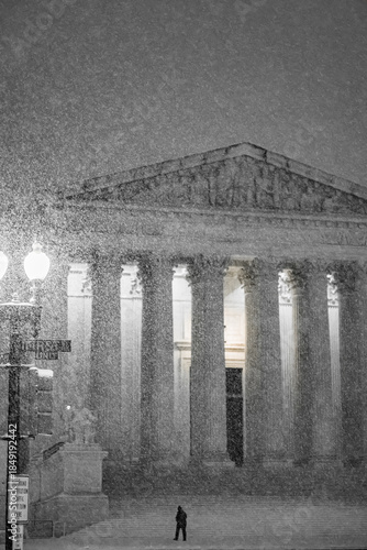 View of a solitary figure braving the blizzard's wrath before the majestic columns of a neoclassical building, bathed in eerie light, Washington DC, United States.
