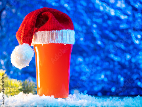 Craft beer glass in Santa's Christmas knitted hat on a snowy blue background, copy space