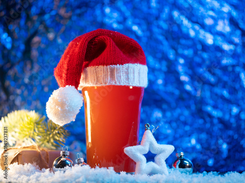 Craft beer glass in Santa's Christmas knitted hat on a snowy blue background, copy space