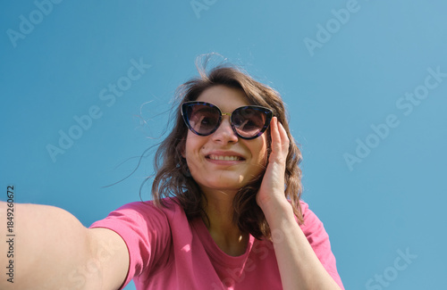 Beautiful carefree young woman in sunglasses and pink t-shirt taking selfie on camera on blue sky background low angle shot.