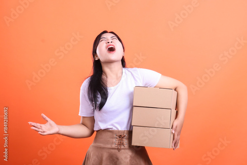 A young woman is visibly stressed while carrying a stack of cardboard boxes, seemingly overwhelmed by the weight and responsibility of delivery. isolated on orange background