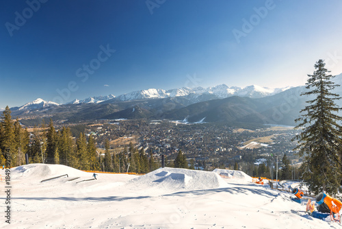 Fototapeta Naklejka Na Ścianę i Meble -  A panoramic view shows a snowy mountain range under a clear blue sky, with a ski slope in the foreground and city afar. Zakopane town at the foot of the Tatra Mountains in Poland, Europe.