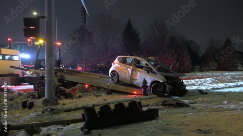 POV damaged compact car being loaded onto a tow truck after a serious accident on a snowy road. Emergency lights illuminate wrecked vehicle, broken traffic pole.
