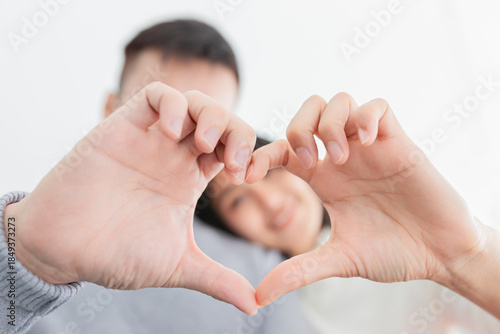 Asian couple making heart shape with hands, sitting close together on sofa. Man and woman in grey and white sweaters showing love gesture. Romantic relationship and affection concept at home.