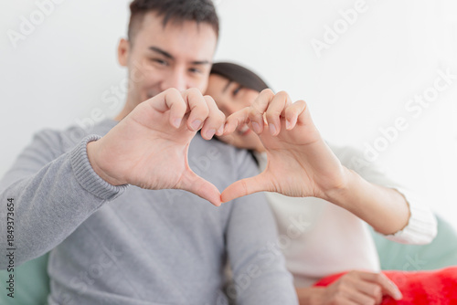 Asian couple making heart shape with hands, sitting close together on sofa. Man and woman in grey and white sweaters showing love gesture. Romantic relationship and affection concept at home.