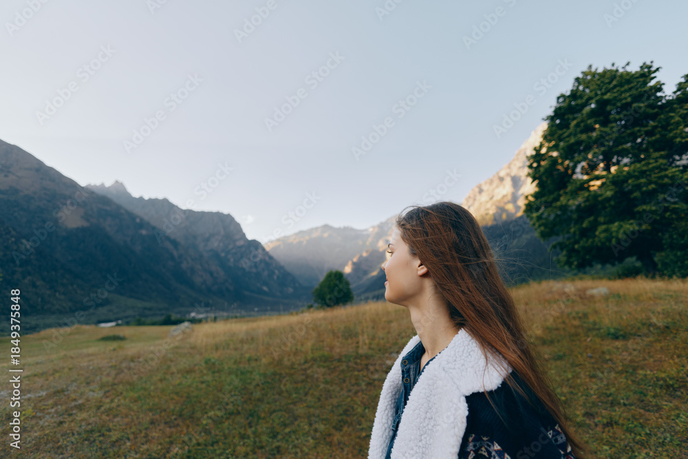 Fototapeta premium Woman in profile looks at distant mountains across a wide meadow landscape, a nature scene outdoors and travel concept with peaceful mood, long hair and layered jacket.