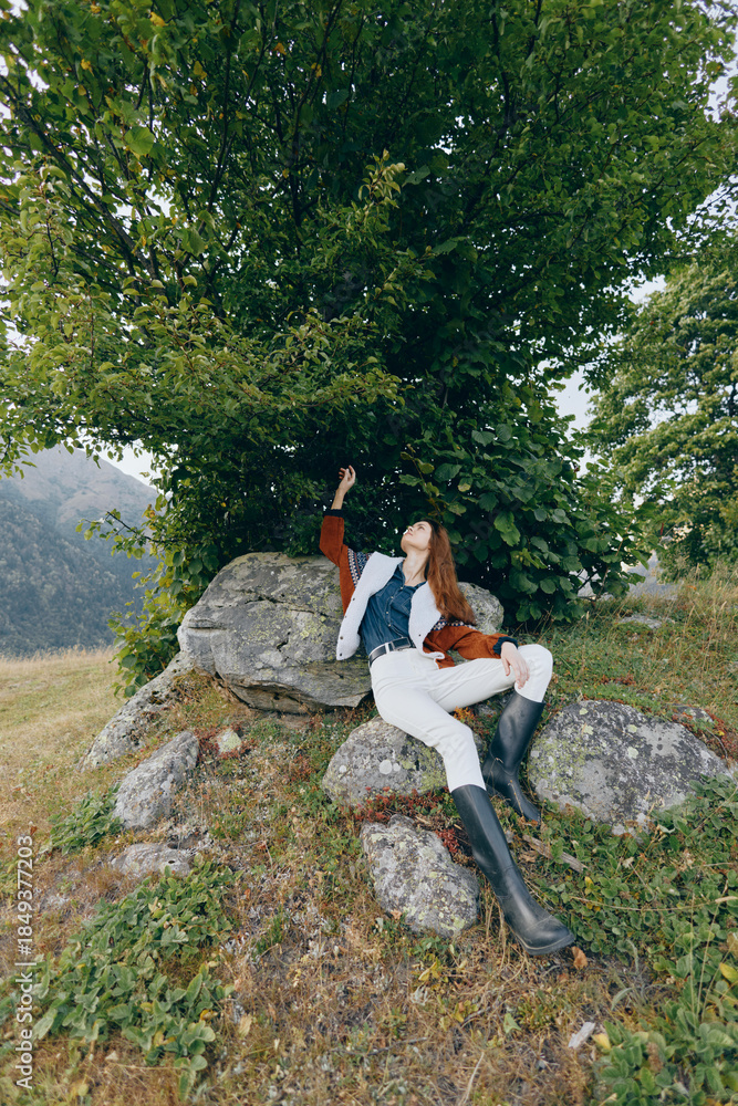 Fototapeta premium Woman relaxing on rocks under a tree in the countryside nature, wearing boots and a casual jacket, enjoying outdoor serenity on a grassy hillside near mountain scenery.
