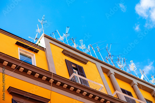Fényképezés Yellow building with white balconies and brown shutters rises beneath blue sky, crowned by chaotic array of rooftop antennas